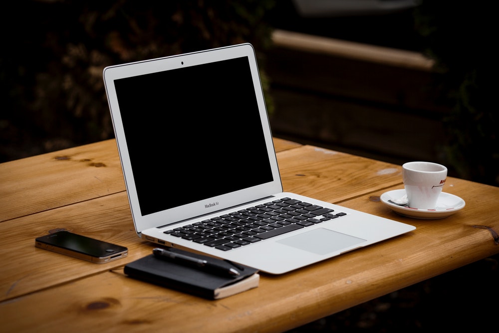 A macbook laptop on a desk with other things like iphone, mug, pen and a notebook